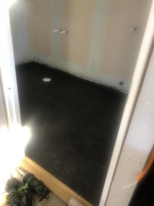 Ett tomt rum under renovering med svart golv, oslipade väggar och byggmaterial i förgrunden.