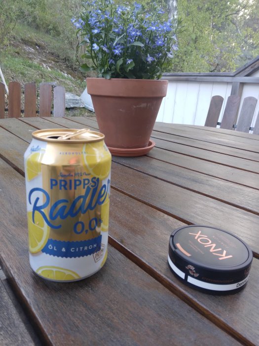 En burk alkoholfri Pripps Radler, snustobaksdosa, blomkruka med blå blommor på ett träbord utomhus.