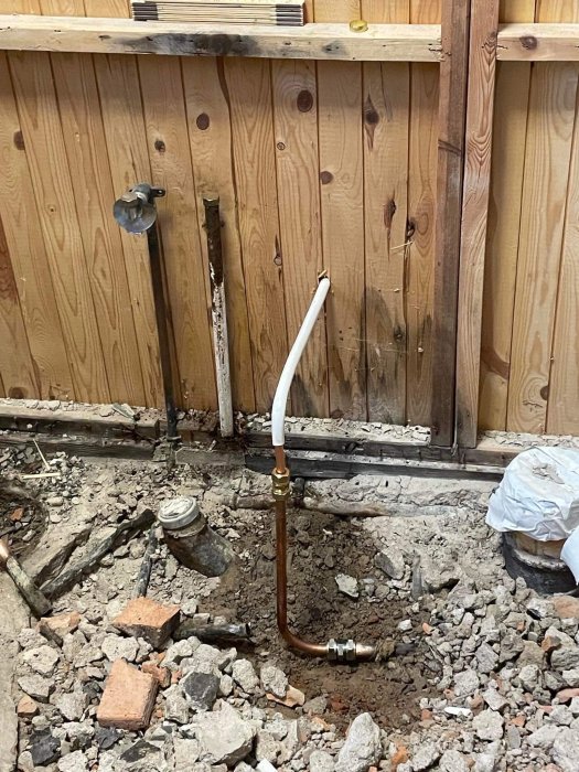 Rörledning och grus på golv vid trädvägg inomhus, möjlig renovering eller reparationsarbete pågår.