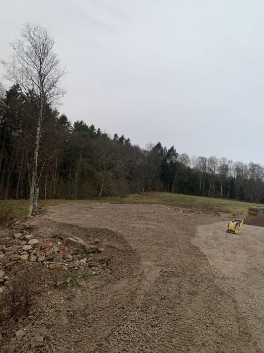 Grusig mark, skogsbakgrund, gul grävmaskin, molnig himmel, hög med sten och tegel, ingen person synlig.