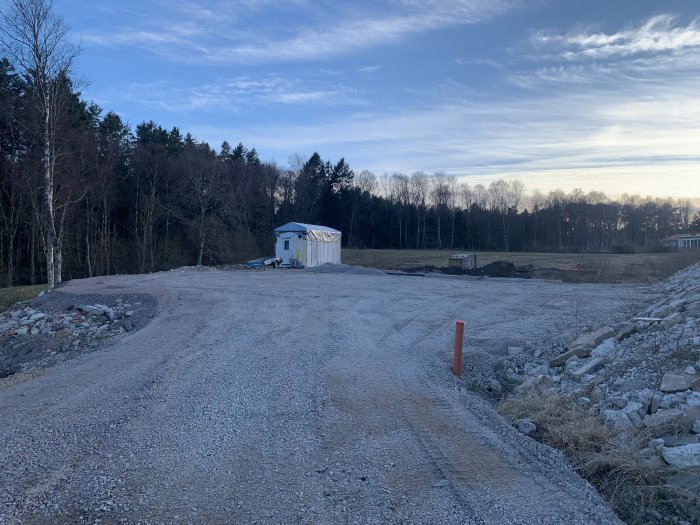 Grusväg leder till byggarbetsplats med skog, tillfällig byggnad och blå himmel i bakgrunden.