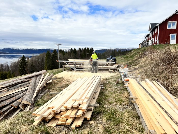 Arbetsplats utomhus med timmer och virke, arbetare i väst, röd byggnad, sjö och snöklädda berg i bakgrunden.