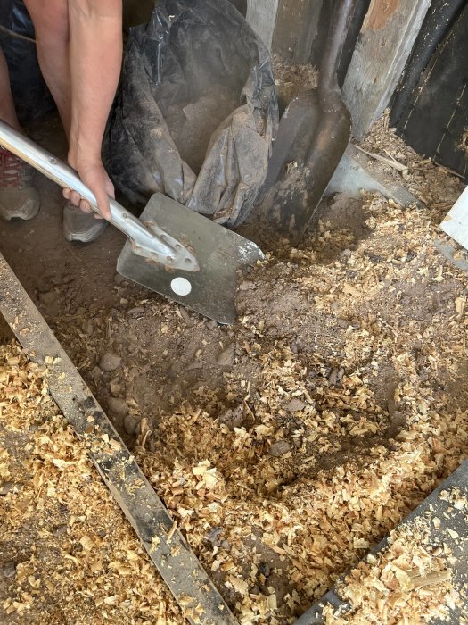 Person städar stall med spade, högar av spån på betonggolv, svart sopsäck, träväggar.