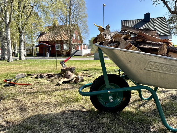 En skottkärra fylld med ved, yxa och röda verktyg på en solig gräsmatta, hus i bakgrunden.