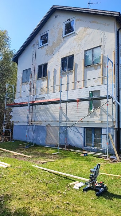 Renovering pågår, hus med byggställningar, gammal färg skrapas bort, såg och brädor på marken.