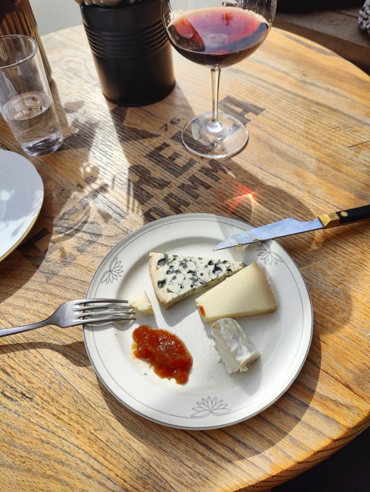 Ostbricka med tre sorter ost och marmelad, bredvid rödvinsglas, på träbord med solljus.