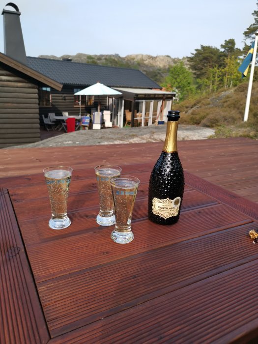 En flaska mousserande vin och tre glas på ett träbord utomhus med byggnad och natur i bakgrunden.