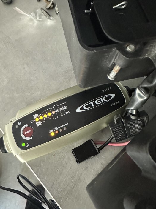 Batteriladdare CTEK MXS 5.0 ansluten, laddningsindikatorer tända, svart bakgrund, kablar synliga.