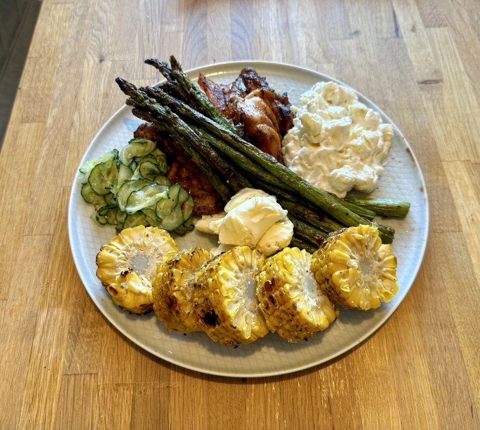 Tallrik med grillad majskolv, sparris, potatissallad, gurksallad och rostad kyckling. Mat på träbord.