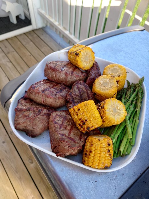 Grillade biffar, majs och sparris på ett serveringsfat utomhus på en träveranda.