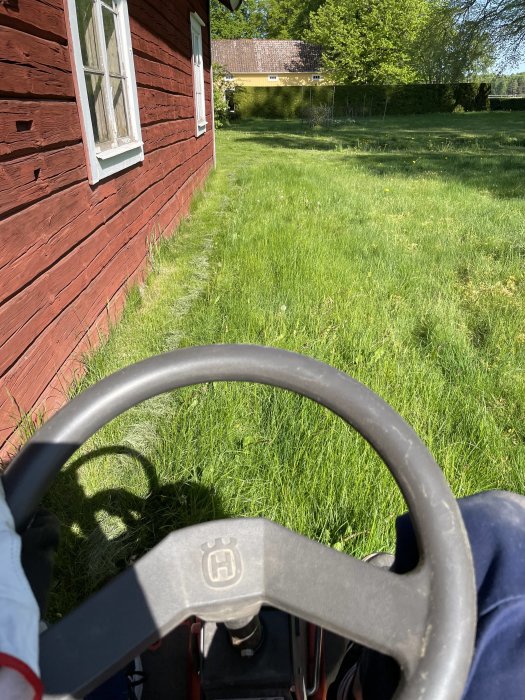 Ratt på gräsklippare, person klipper gräs, röd trästuga, grönt gräs, soligt, utomhus.