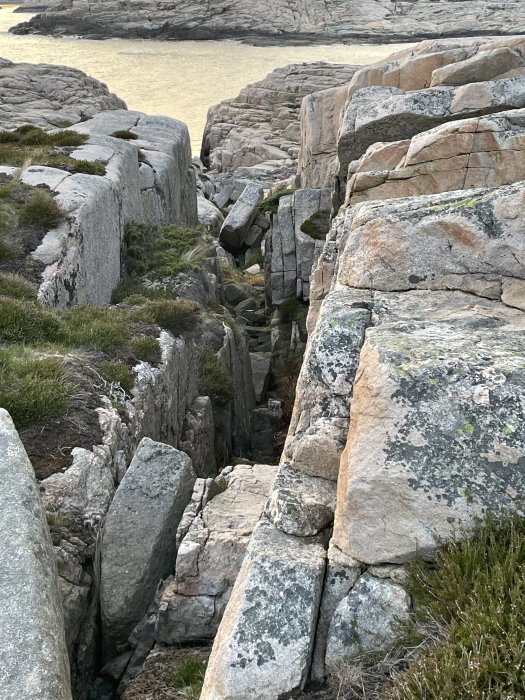 Klippor med sprickor nära vatten, grönt gräs, ljus himmel, naturlandskap.