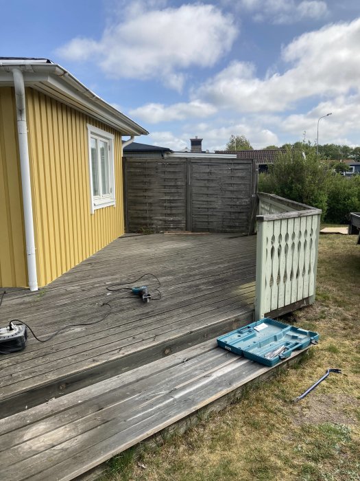 Trädäck vid gult hus, verktyg utspridda på marken, molnig himmel, staket, renoveringsarbete pågår.