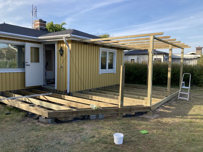 Ett hus med pågående byggnation av en träterrass med pergola, verktyg synliga, klart väder.