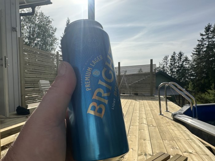 Person håller en ölburk utomhus, soligt väder, träaltan, pool och träd i bakgrunden.