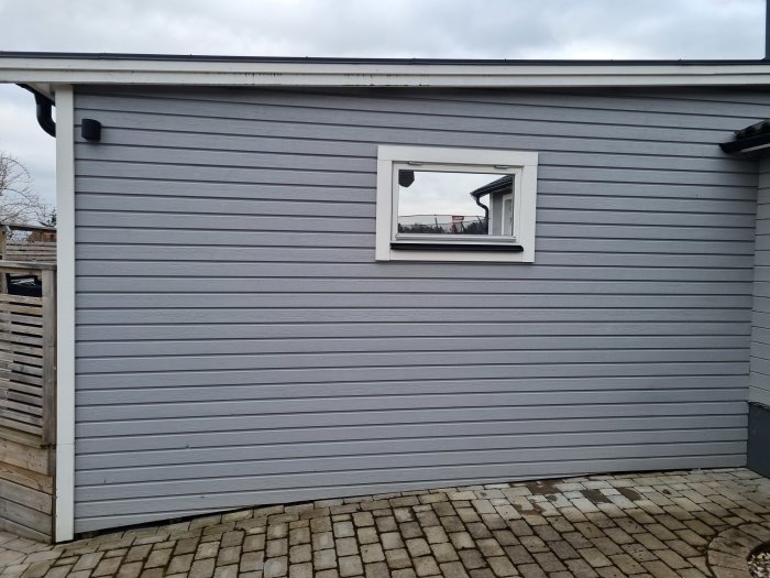 Grått hus med fönster, stenläggning framför, reflektion i fönstret, molnig himmel överst.