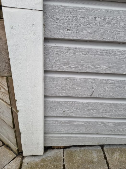 Vit yttervägg i trä, vertikala och horisontella plankor, grå stenplattor på marken.