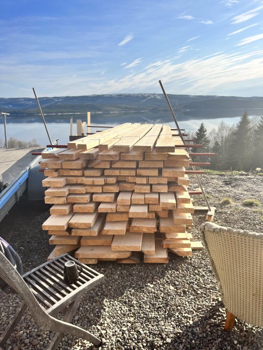 Stapel virke vid byggarbetsplats med utsikt över sjö och berg. Soligt väder, klarblå himmel.