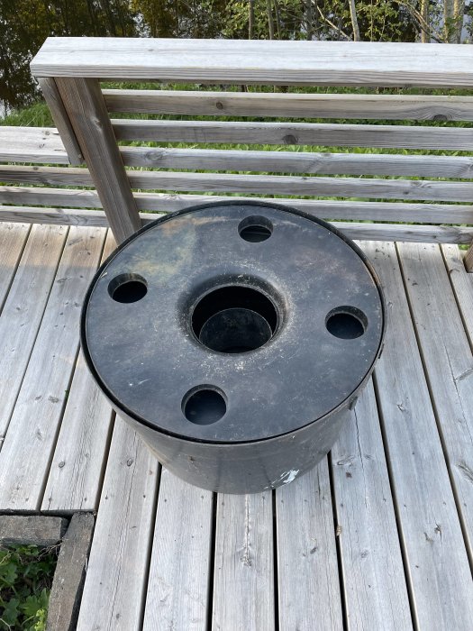 Svart askkopp på trädäck, runda hål, bredvid bänk, utomhusmiljö, ingen synlig rök eller aska.