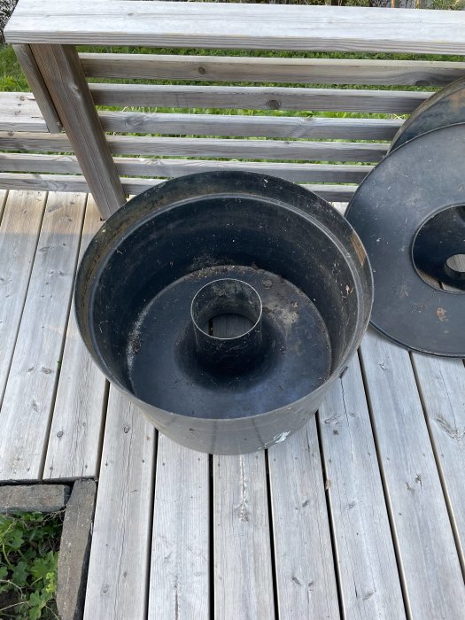 Svart sopbehållare utan lock på trätrall med räcke i bakgrunden.