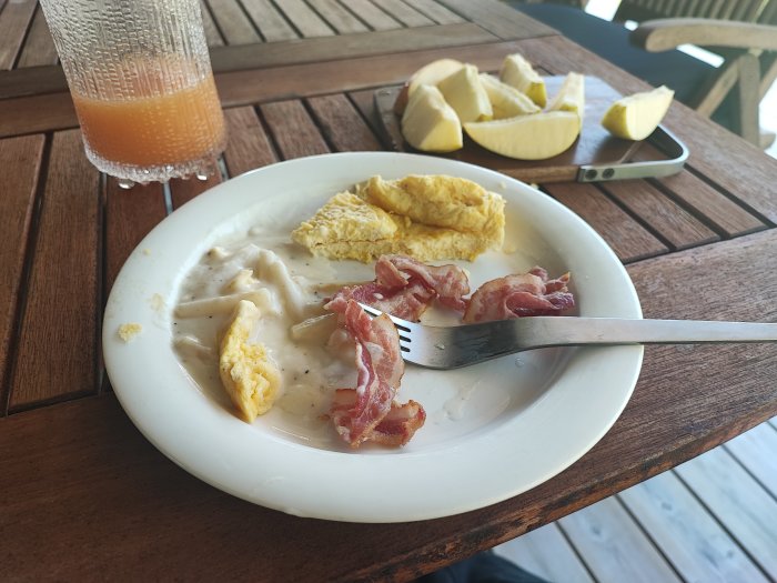 Halväten frukost med bacon, ägg, sås, appelbitar, juice, och en smartphone på träbord.