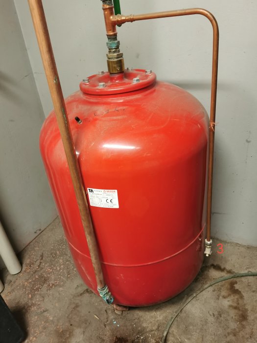 Röd expansionskärl för värmesystem med kopparledningar och tryckmätare i källare.