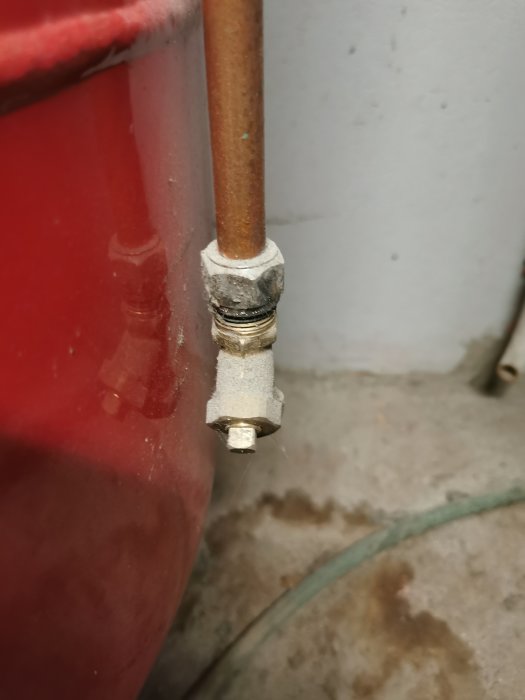 Kopparledning ansluten till röd behållare genom koppling med mutter. Betonggolv och vit vägg bakgrund.