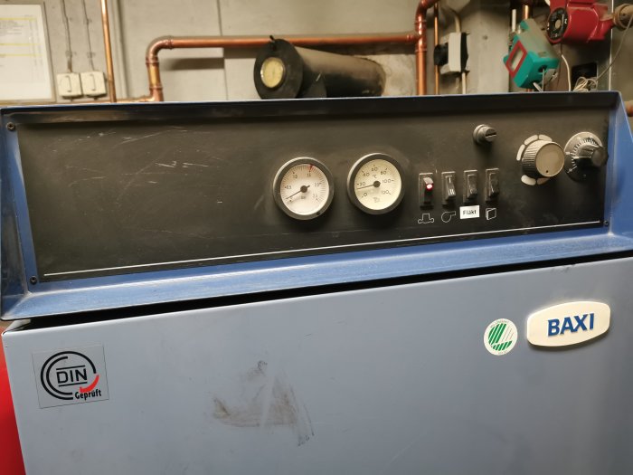 Blått kontrollpanel med mätare, knappar och reglage, märken ”DIN” och ”BAXI”, rörlagning i bakgrunden.