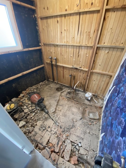 Rivningsarbete inomhus, exponerade rör, trasigt tegelgolv, verktyg, träväggpanel, renoveringsprojekt pågår.