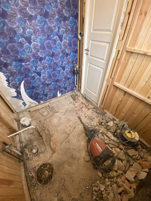 Renoveringsarbete pågår, rivet golv och vägg, byggverktyg och kablar synliga, blommigt tapet i bakgrund.