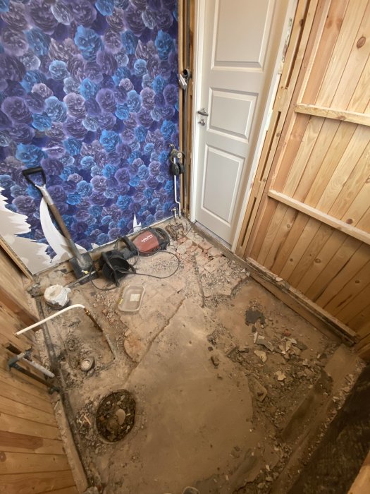 Renovering pågår, rörledningar synliga, rivet golv, vägg med blommönster, verktyg och byggavfall.