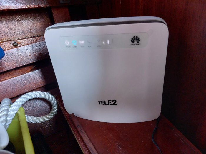 En Huawei-router märkt Tele2. Indikatorlampor lyser. Står på mörkt trä. Kabel och rep i bakgrunden.