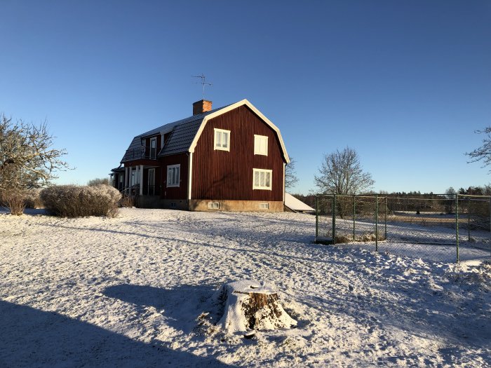 Rött svenskt hus med vita knutar, snötäckt mark, klart väder, vinterdag, trädstubbe, staket, blå himmel.