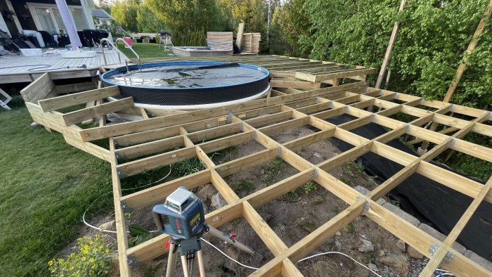 Trädäck under konstruktion, trampolin, grönska, byggarbetsplats med verktyg, utomhus i ett bostadsområde.
