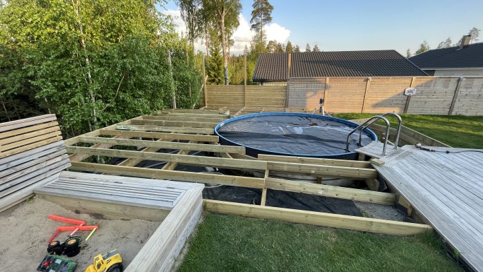 Trädgård med pågående bygge av däck runt trampolin, leksaker på sand, trästaket i bakgrund.