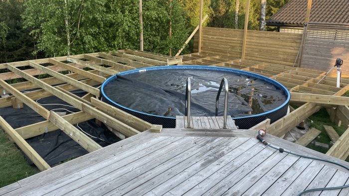 Utomhuspool omgiven av oavslutad trätralldäck, grönska i bakgrunden, skymning.