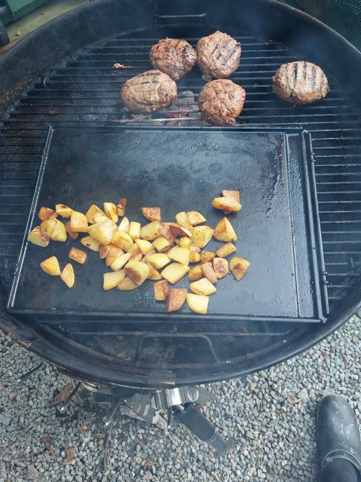 Grill med hamburgare och klyftpotatis, matlagning utomhus, svart grill, grusunderlag, grillverktyg på marken, matlagningsaktivitet.