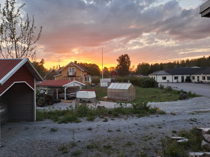 Lantlig vy, solnedgång, bostadshus, växthus, fordon, trädgård, moln, skymning, lugn atmosfär.