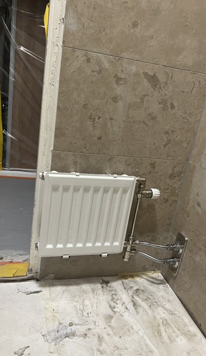 Vit radiator på vägg, ansluten med rör, oavslutad byggnadsplats, öppen dörr, stenplattor, dammigt golv.