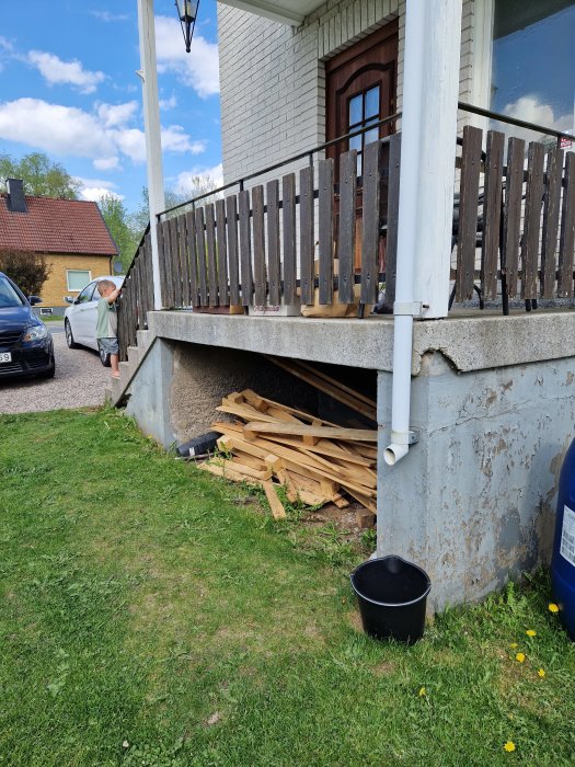 Ett hus med veranda, barn vid staket, träbrädor under verandan, bil, gräs och hink.