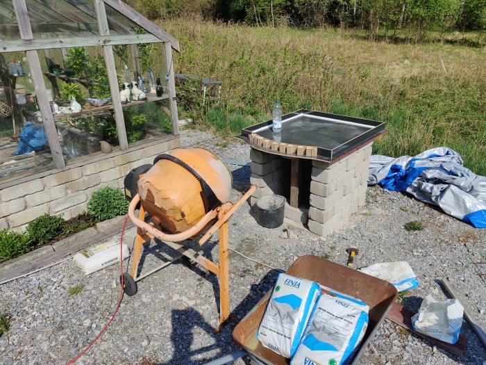 Utomhusarbetsplats med cementblandare, oavslutad ugn, säckar med byggmaterial och drivhus i bakgrunden.