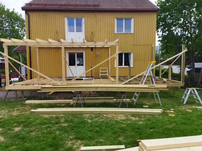 Träkonstruktion framför gult hus, byggarbetsplats, virke, stegar, gräsmatta, pågående utbyggnadsprojekt.