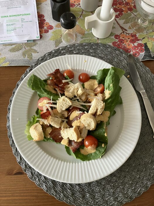 Caesarsallad med bacon, krutonger, körsbärstomater på vit tallrik, kryddkvarnar och mortel i bakgrunden.