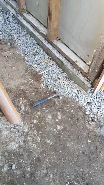 Byggarbetsplats med grus, jord, träbitar och en hammare på marken vid en väggkonstruktion.