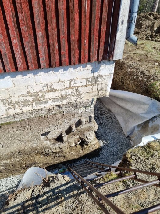 Rödfärgad stuga, vit grund, erosionsskador, reparation pågår, grus, armeringsjärn, grävschakt, skyddsplast, soligt väder.