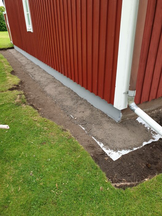 Nylagd trottoarkant vid hus, grön gräsmatta, röd vägg, stuprör, nypreparerad jord.