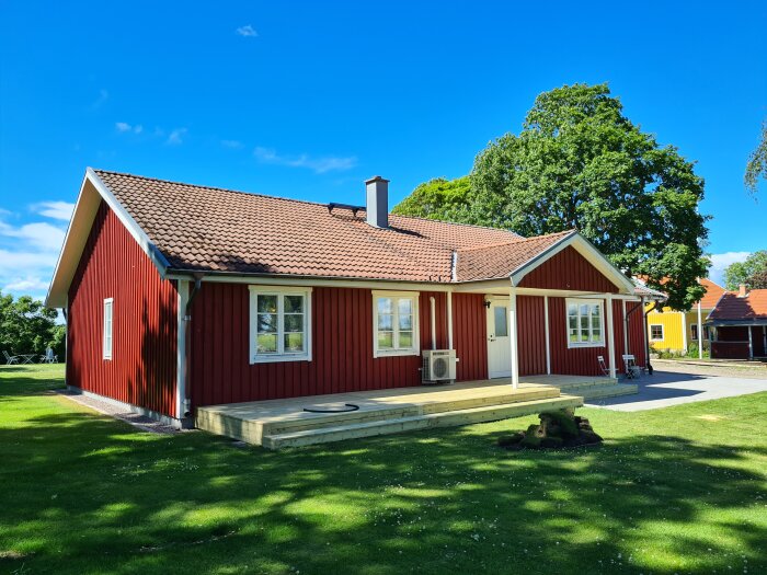 Traditionellt rött hus med vita knutar, trädäck, grönt gräs och klarblå himmel.