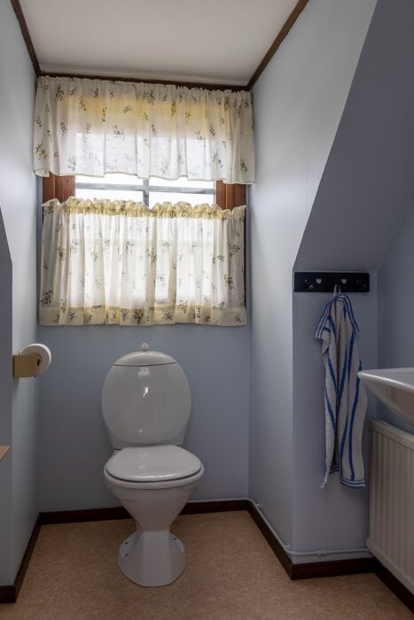 En liten toalett, vita väggar, blommiga gardiner, beige golv, handduk, toalettpapper och en toalettstol.
