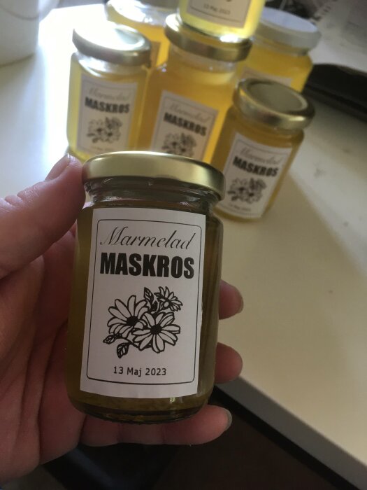 Hand håller litet glasburk märkt "Marmelad Maskros", bäst före datum och fler burkar i bakgrunden.