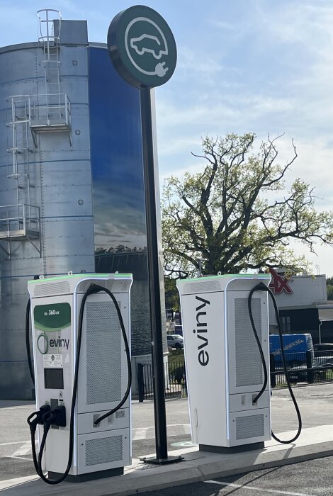 Två elbils laddstationer, skylt med elbilssymbol, silo och träd i bakgrunden.